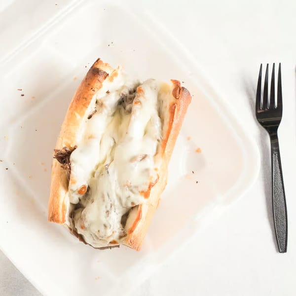 A cheesy steak sub sandwich with melted white cheese and meat on a white plate with a fork.