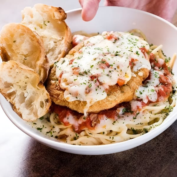 Chicken parmesan with spaghetti, marinara, melted cheese, and garlic bread in a white bowl.